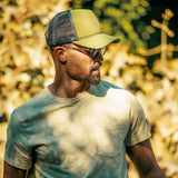 Man in mesh trucker hat and sunglasses wearing beige t-shirt outdoors in sunlight