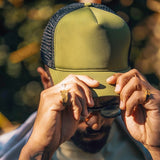 Man wearing olive green mesh trucker cap outdoors, adjusting brim with both hands