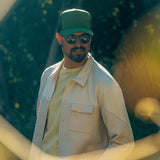 Man outdoors wearing a light quilted jacket, green cap, and sunglasses in sunlight