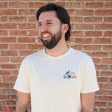 Man with dark hair and beard wearing a short-sleeve cotton graphic tee stands before a red brick wall.