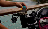 A cyclist secures a metal mug into a black tube-shaped caddy on the handlebar by a river, highlighting its NocLoc fastener.