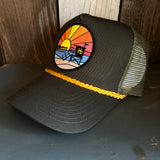 Mesh trucker hat with sunset beach patch and yellow braided trim on dark wood background