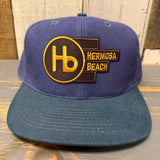 Navy and dark green 6-panel low profile baseball cap with yellow Hb logo, displayed on a wood background.