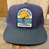 Navy and dark green 6-panel cap with adjustable strap and surfer patch, suitable for casual outdoor wear.