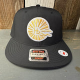 Black snapback hat with embroidered yellow sunburst and HB logo on a wood background