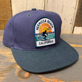 Navy and dark green cap with a patch featuring a surfer, sun, and pier; suitable for casual outdoor wear.