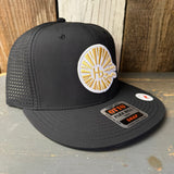 Black mesh snapback hat with gold sun and waves patch, displayed on rustic wood surface
