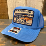 High crown blue trucker hat with a patch featuring a pier and text on a wooden surface.