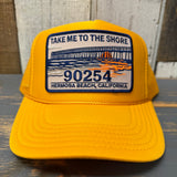 Gold trucker hat with a patch featuring 'Take Me to the Shore' and 'Hermosa Beach, California' text on a wooden surface.