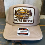 Khaki trucker hat with a graphic of a van and mountains, featuring the text 'You're Trippin' on a wooden surface.