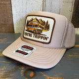 Khaki trucker hat with a you're trippin patch and Otto sticker on a wooden surface.