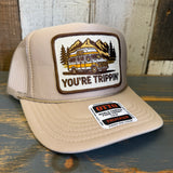 Khaki trucker hat with a graphic of a van and mountains, text 'You're Trippin' on a wooden surface.