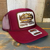 Maroon trucker hat with 'You're Trippin'' graphic and van design on a wooden surface.