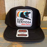 Black trucker cap with 'Surf Hermosa' logo on a wooden surface.