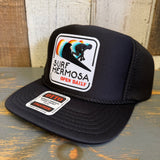 Black trucker hat with a surf-themed logo on a wooden surface.