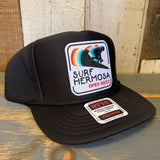 Black trucker hat with 'Surf Hermosa' logo on a wooden surface.