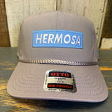 Gray cap with 'Hermosa' patch and 'Otto' label on a wooden surface