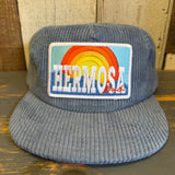 Slate blue corduroy cap with a colorful patch featuring 'Hermosa Beach' on a wooden surface.
