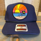 Navy blue cap with a colorful beach scene patch on a wooden surface.