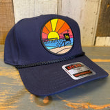 Navy blue cap with a colorful beach scene patch on a wooden surface.