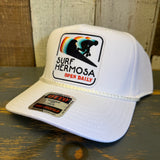 White cap with 'Surf Hermosa' logo on a wooden surface.