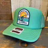 Cactus green trucker hat with a surfing patch, mesh back, and foam front rests on a wooden surface.