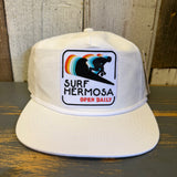 White cotton hat with a colorful surfer patch and “Surf Hermosa Open Daily” text on the front panel.