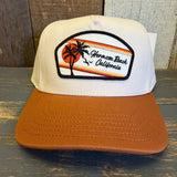 white canvas trucker hat with a black and white 'Hermosa Beach California' patch and a mustard brown brim on a wooden surface
