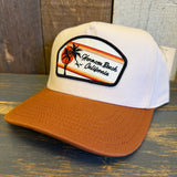 white canvas trucker hat with a black and white 'Hermosa Beach California' patch and a mustard brown brim on a wooden surface