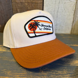 white canvas trucker hat with a black and white 'Hermosa Beach California' patch and a mustard brown brim on a wooden surface