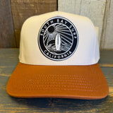 white canvas trucker hat with a black and white 'South Bay Surf California' patch and a mustard brown brim on a wooden surface