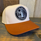 white canvas trucker hat with a black and white 'South Bay Surf California' patch and a mustard brown brim on a wooden surface