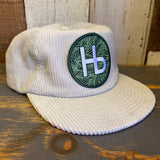 white corduroy cap with a green and white HB patch on a wooden surface