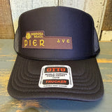 Black high crown trucker hat with curved brim, shown on a wooden surface; mesh panels and structured front visible.