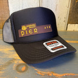 Black high-crown trucker hat with curved brim, mesh back, and PIER AVE patch displayed on wooden surface.