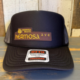 Black high crown trucker hat with patch and sticker on curved brim, mesh back, suitable for casual outdoor wear.