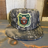 Surf camo SMOKEY BEAR 5-panel hat with gold border, displayed on rustic wood. Cotton material, casual outdoor use.