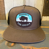 Khaki and black bison graphic 5-panel trucker hat with mesh back, shown on rustic wooden surface.