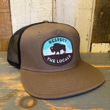 Khaki and black 5-panel trucker hat with bison graphic, mesh back, and snapback closure on a wooden surface.
