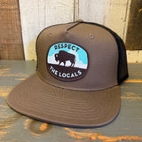 Khaki and black 5-panel trucker hat with bison graphic, mesh back, displayed on a rustic wooden surface.