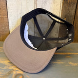 Khaki and black 5-panel trucker hat with mesh back is upside down on a wooden surface.