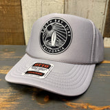 Grey high crown trucker hat with black patch, mesh back, displayed on a wooden surface.