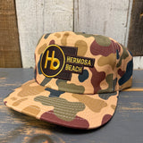 Camo 5-panel hat with yellow patch, made for workwear, displayed on a wooden surface.