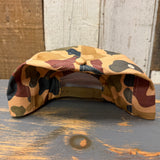 Camo 5-panel work hat, canvas material, displayed upside down on a wooden table with a wood-paneled wall background.