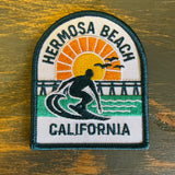 Embroidered patch showing a surfer, pier, sunset, and bird. Multicolor fabric, for jackets or bags.