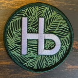 Round green embroidered patch with white Hb lettering on a leafy background, suitable for clothing or accessories.