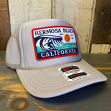 Grey high crown trucker hat with mesh back, displayed on a wooden surface. Suitable for casual outdoor wear.