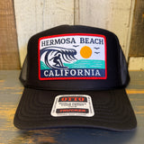 Black high crown trucker hat with front patch showing surfer, wave, sun, and birds; mesh back for breathable wear.