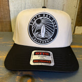 Black and natural 5-panel surf cap with black patch, displayed on a wooden surface. Suitable for casual or outdoor use.
