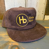 Java brown corduroy hat with yellow logo and brass clasp, displayed on a wooden surface.
